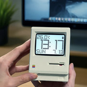 Retro Macintosh Clock held in hands, vintage Macintosh-inspired digital alarm clock displaying temperature, date, and time against a blurred computer screen background